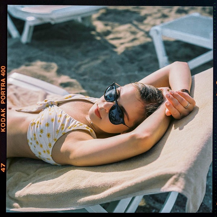A woman wearing sunglasses and a floral bikini top is lying on a lounge chair on a sandy beach.