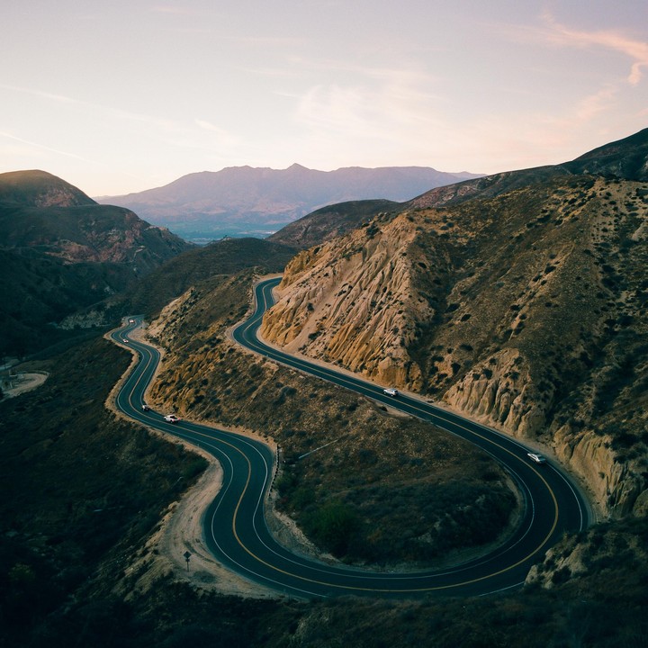 A winding road snakes through a canyon with mountains in the background at dusk.