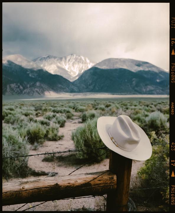 A cowboy hat rests on a wooden fence post with barbed wire, overlooking a vast landscape of brush and snow-capped mountains under a cloudy sky.