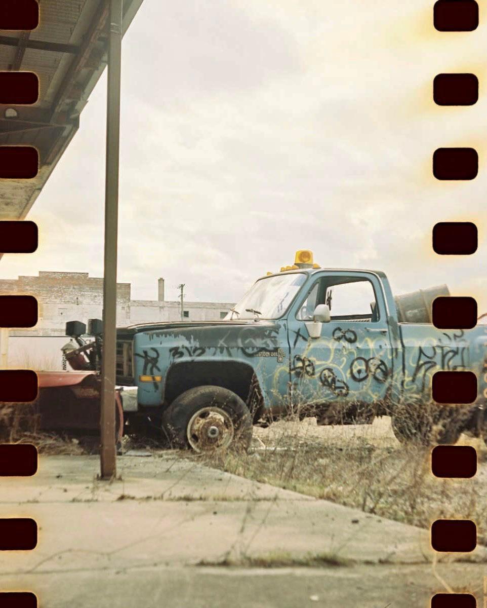 A blue pickup truck with graffiti on its side is parked in a grassy area next to a building.