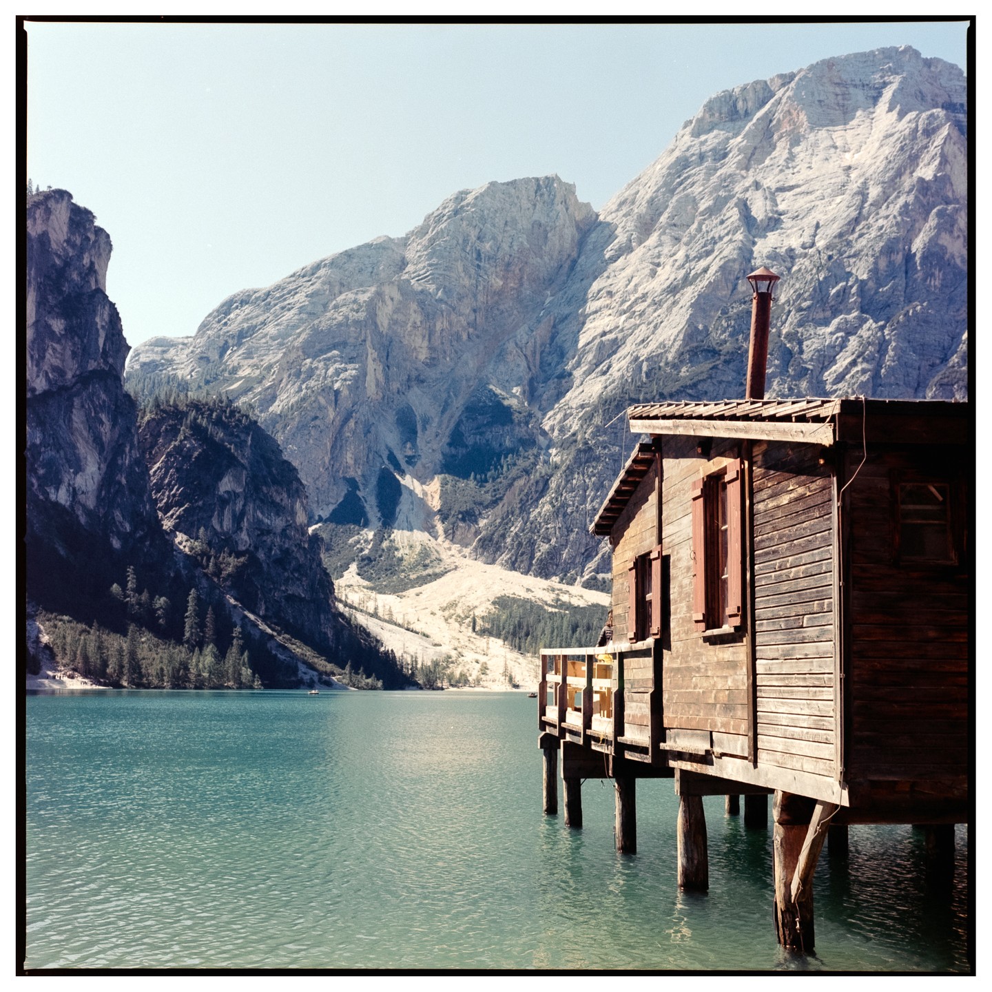 A wooden chalet on stilts sits on a lake with mountains in the background.