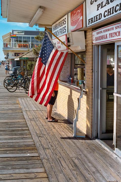 A person stands partially obscured by an American flag on a wooden boardwalk next to a food stand.
