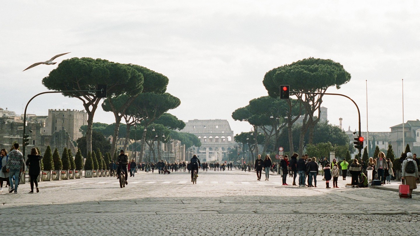 A wide street in Rome is filled with pedestrians and a few cyclists, with the Colosseum visible in the distance and a seagull flying overhead.