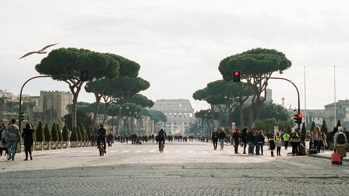 A wide street in Rome is filled with pedestrians and a few cyclists, with the Colosseum visible in the distance and a seagull flying overhead.