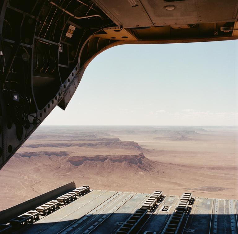 A view from the back of an aircraft shows a vast desert landscape with mesas and canyons under a clear sky.