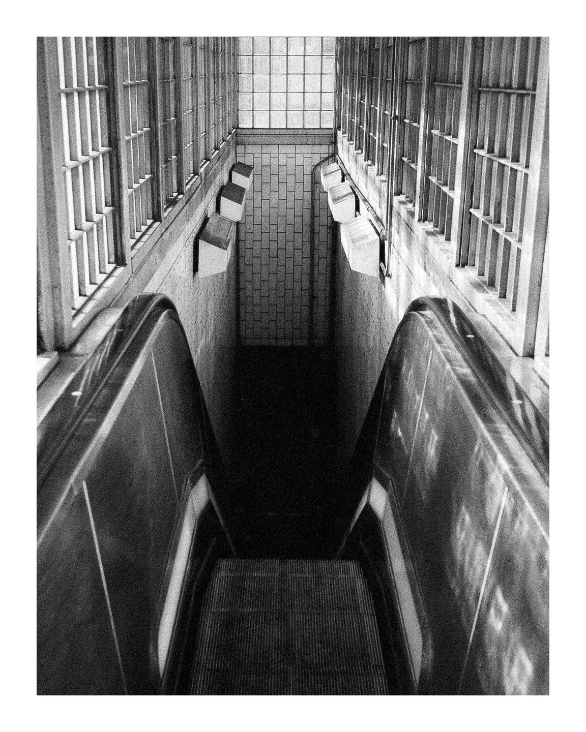 An empty escalator descends into a dark subway station with tiled walls and barred windows.