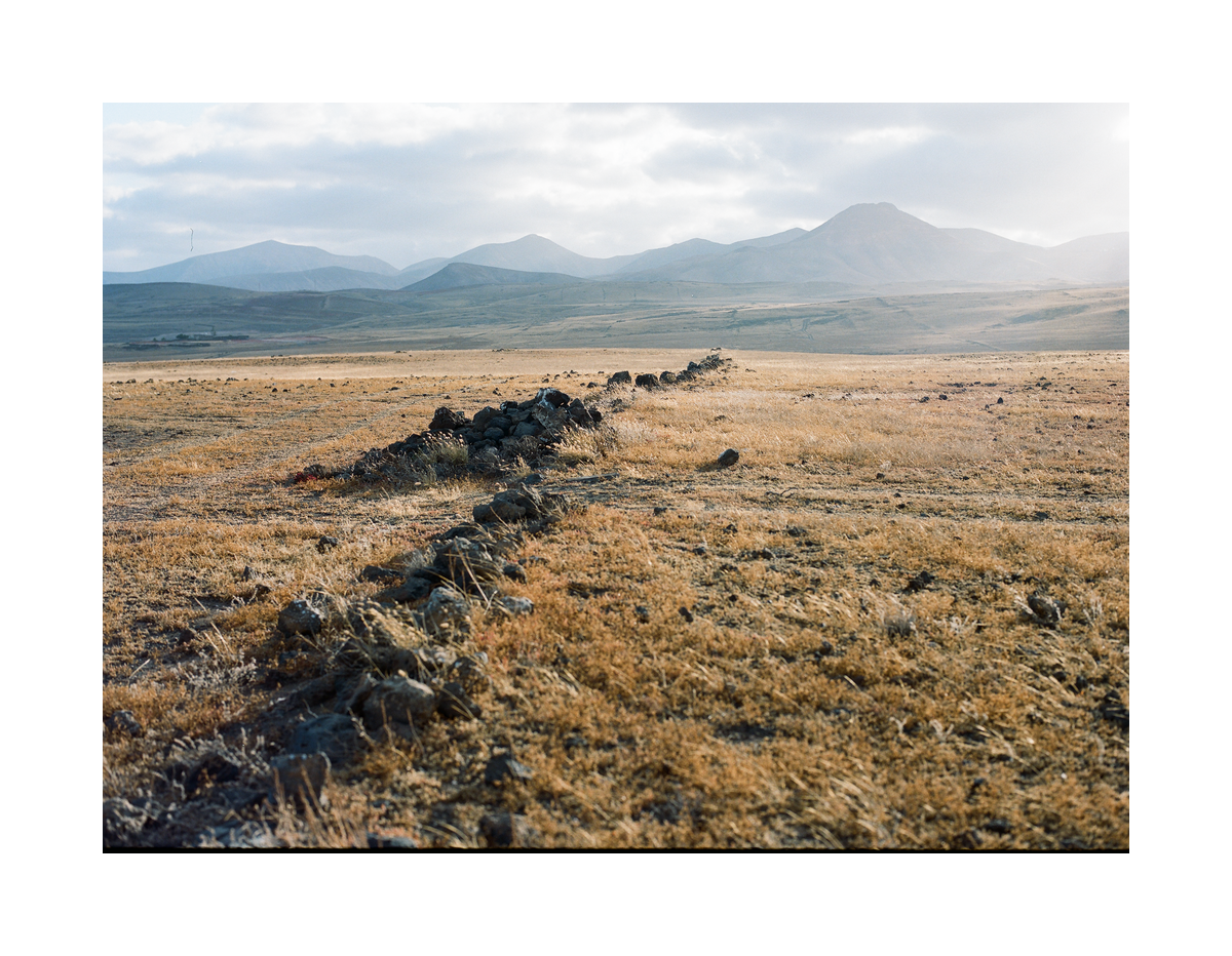 A low stone wall stretches across a field of dry grass toward distant mountains under a hazy sky.