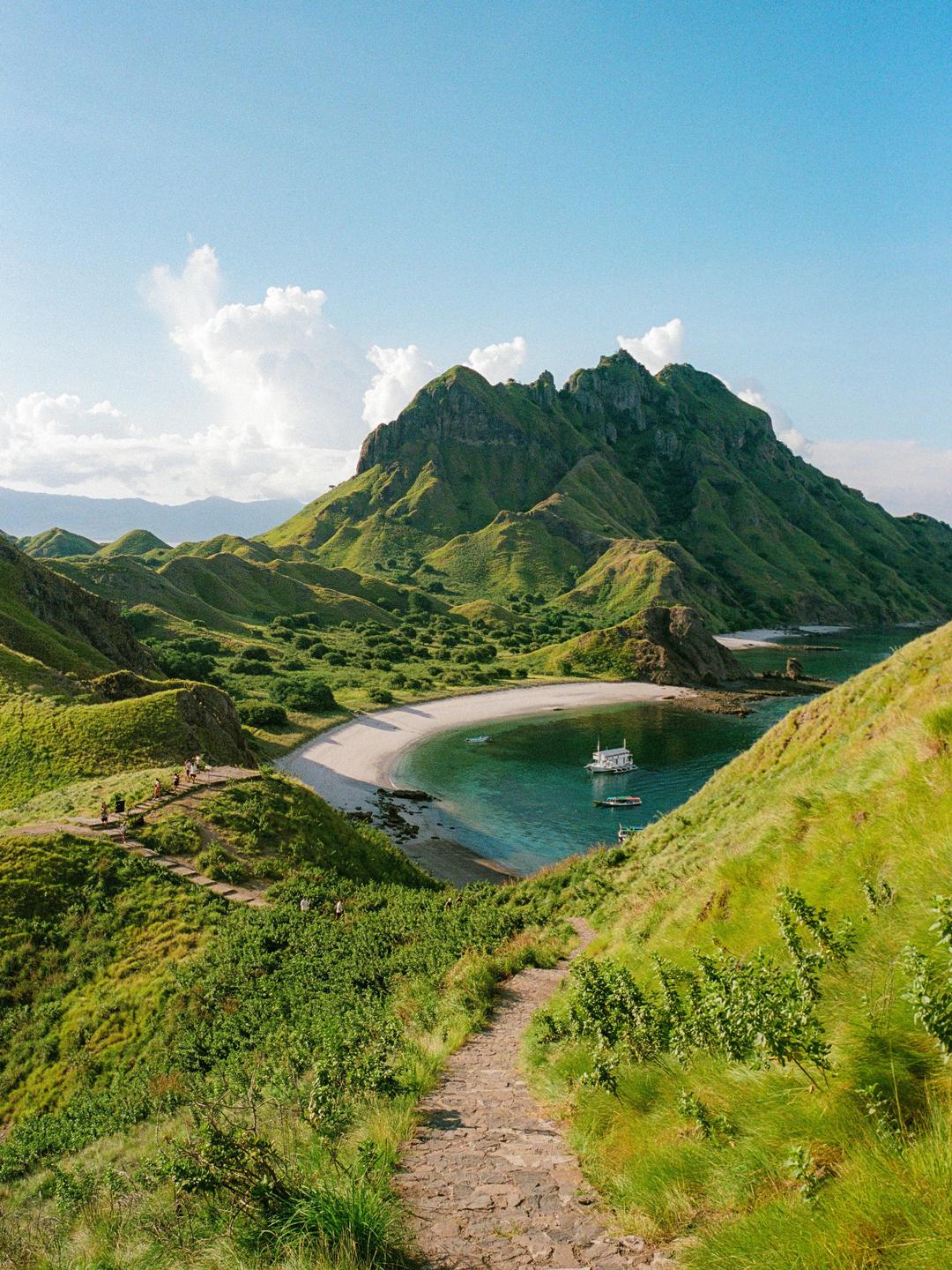 A winding stone path leads down a grassy hillside towards a crescent-shaped beach with boats in the clear blue water, all framed by a large green mountain under a bright sky.