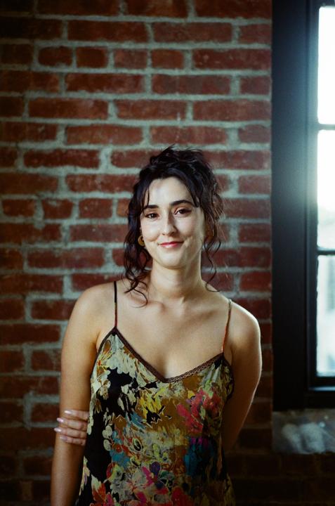 A woman with dark, curly hair smiles at the viewer while standing in front of a brick wall next to a window.