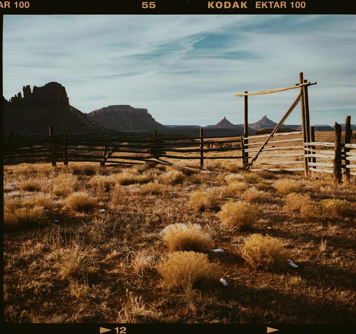 A wooden fence stands in a field of dry brush with mountains in the background under a cloudy sky.