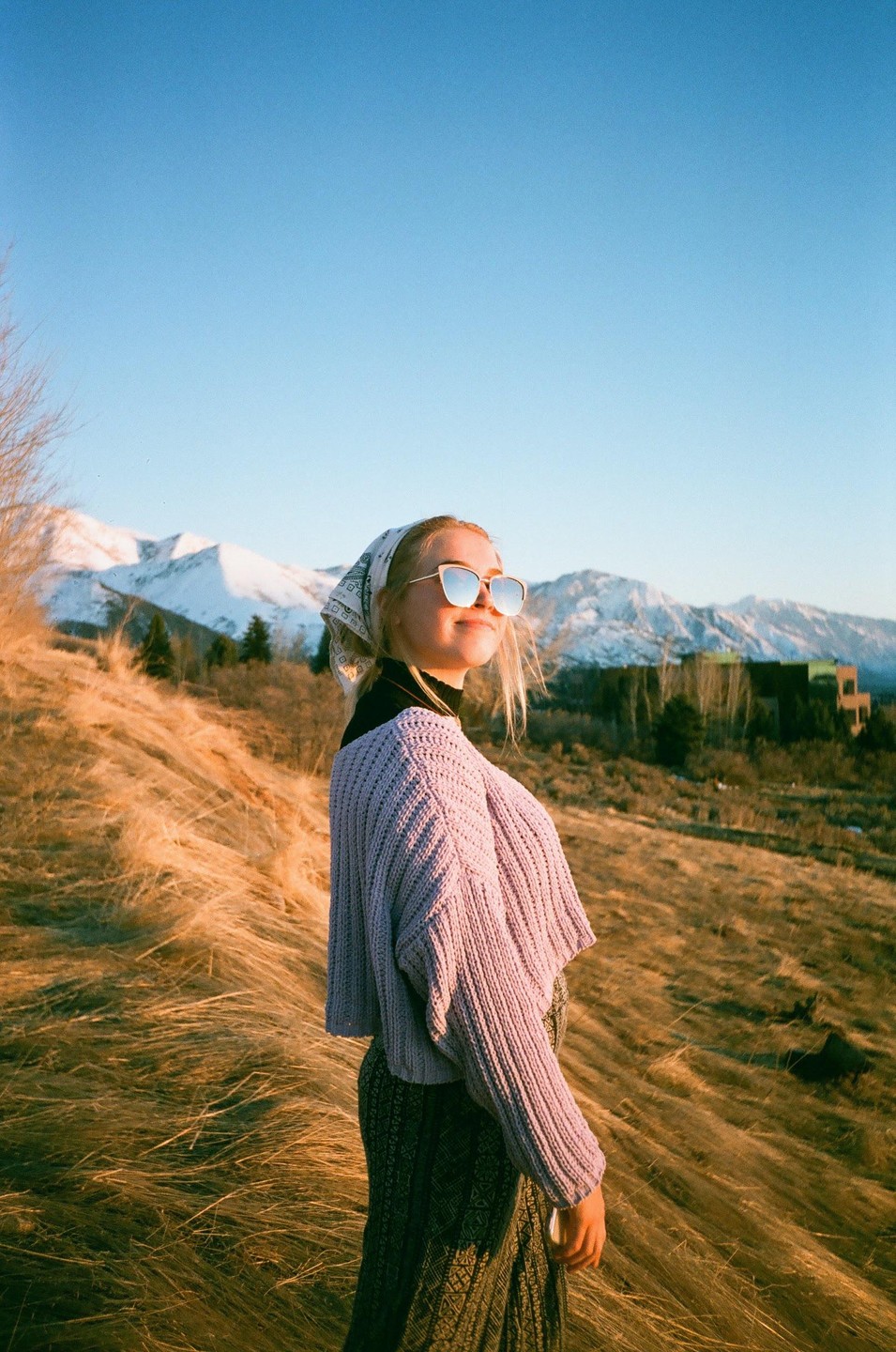 A woman wearing sunglasses and a headscarf stands on a grassy hill with snow-capped mountains in the background.