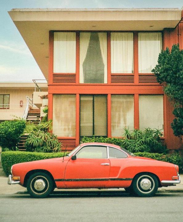 A red car is parked on the street in front of a red building with large windows.