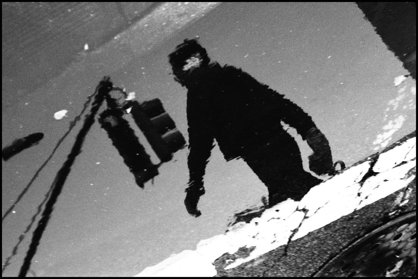 A person and a traffic light are reflected in a puddle on a street in New York.