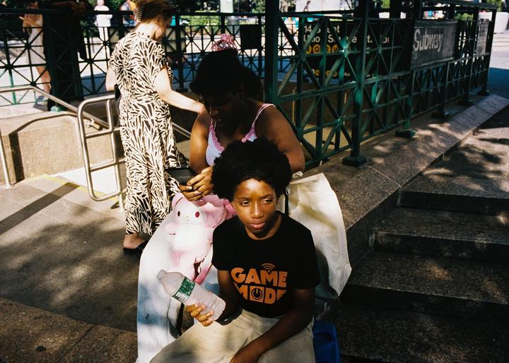 A woman and a boy sit on a street next to a subway entrance while another woman walks past them.