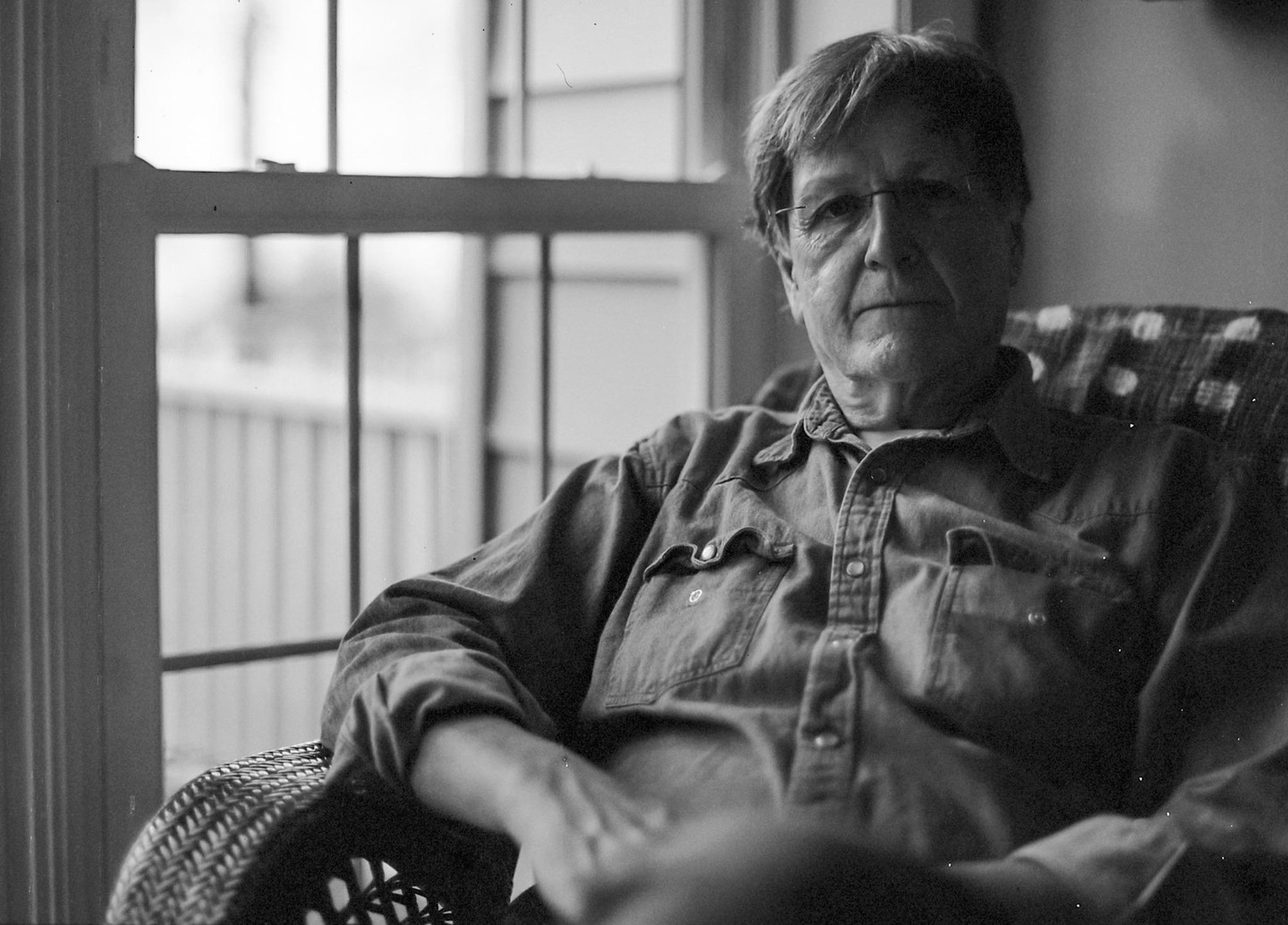 A man with glasses sits in a wicker chair next to a window.