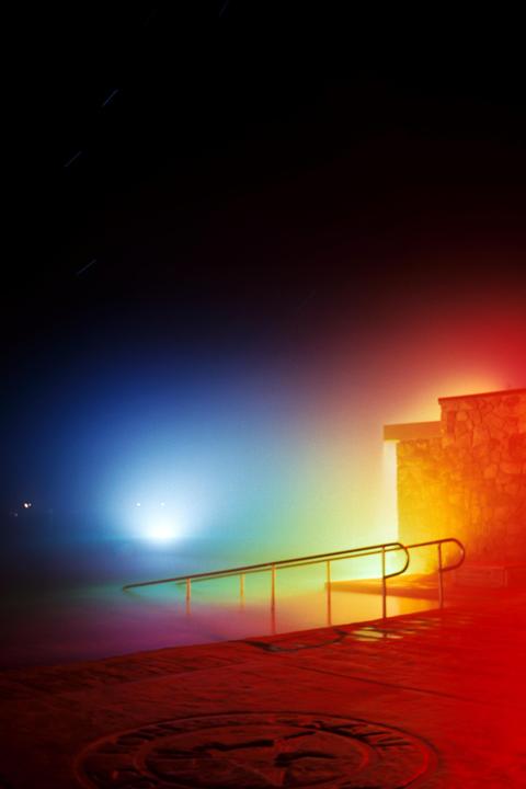 A brightly lit swimming pool with a stone building and railing is visible at night.