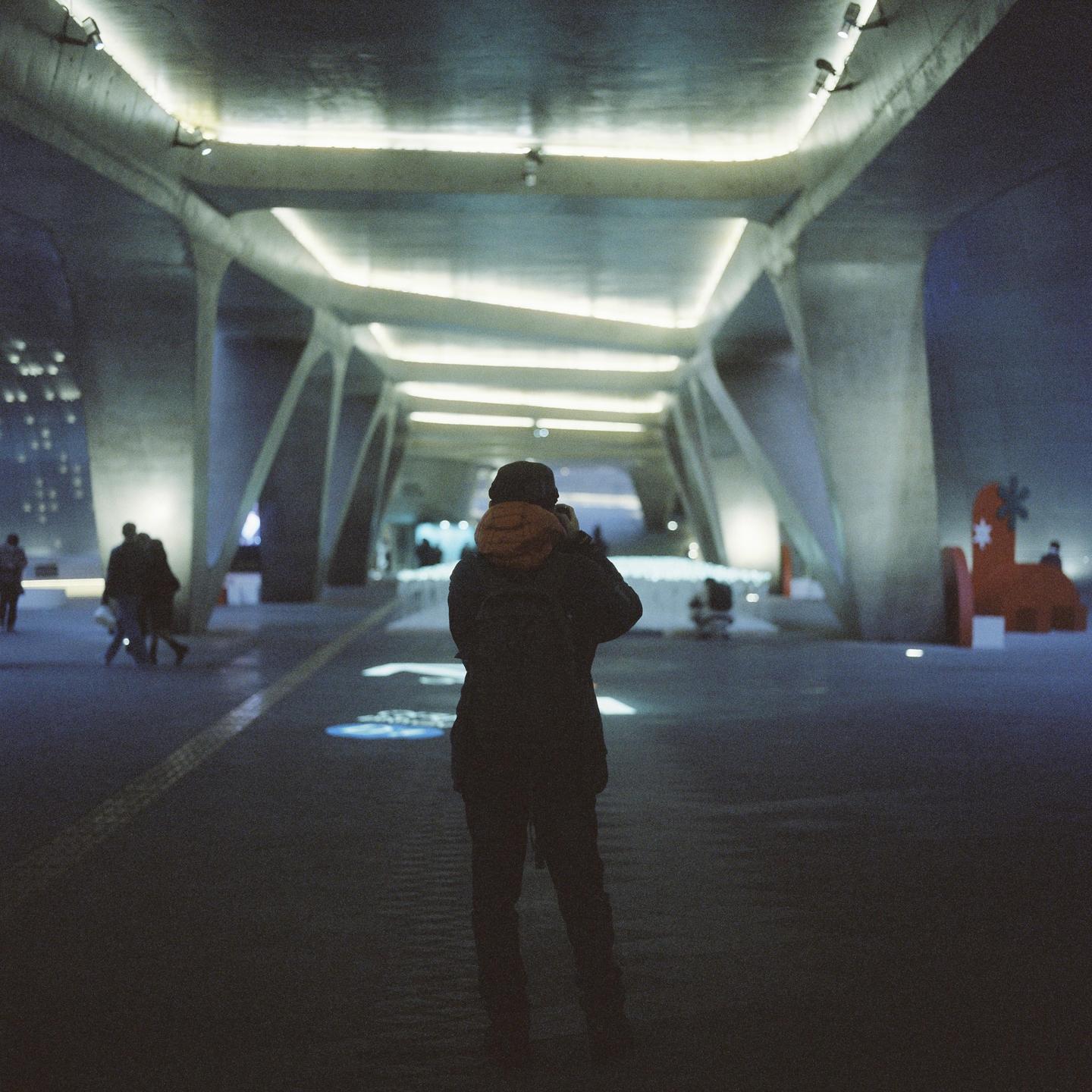 A person stands with their back to the camera, looking down a brightly lit, modern, concrete tunnel at night.