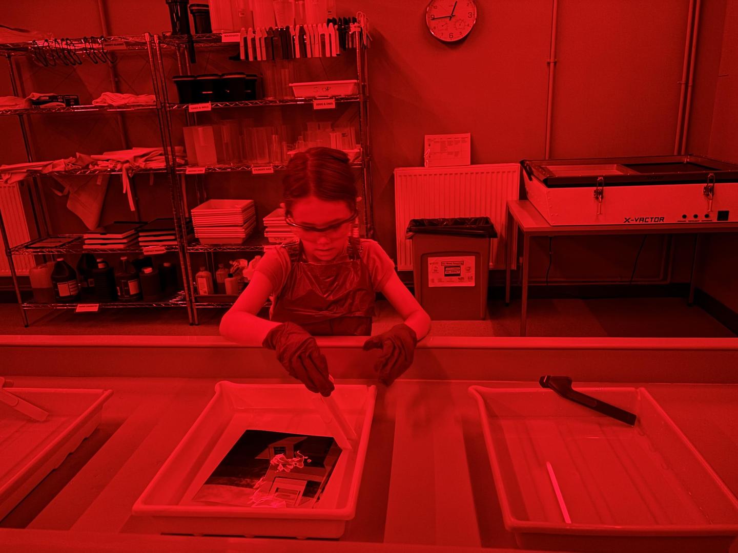 A child wearing safety glasses and gloves is developing a photograph in a darkroom.