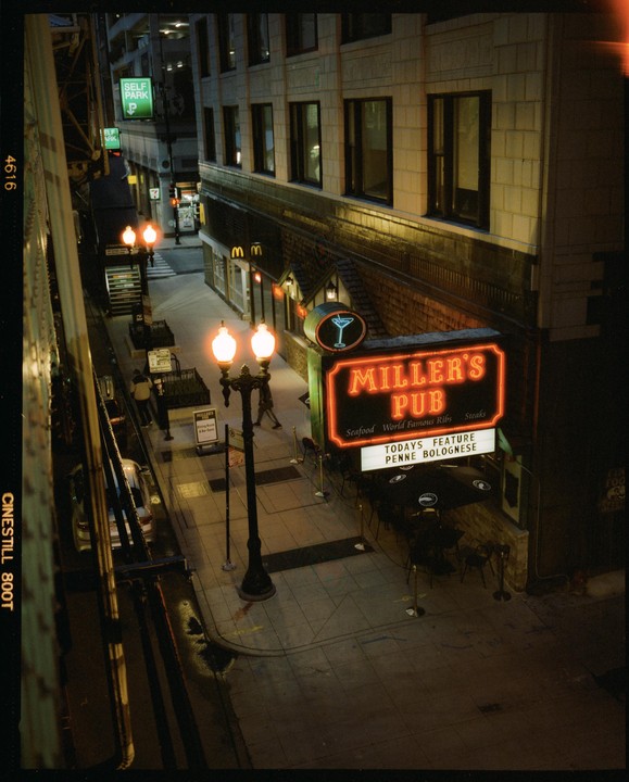 A neon sign for Miller's Pub illuminates a city street at night, with a few people walking on the sidewalk.