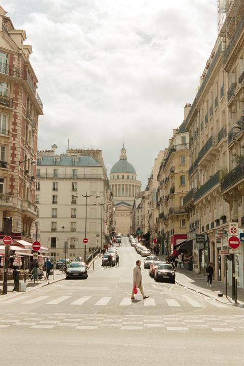A man crosses a street with cars parked on both sides, leading up to a large domed building.