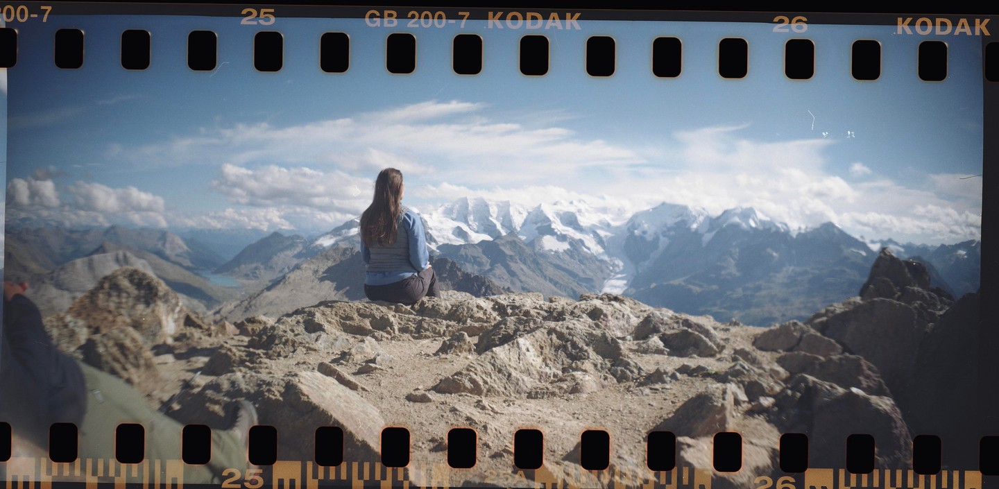 A woman sits on a rocky mountain peak, looking out at a vast landscape of snow-capped mountains and a valley with a lake.