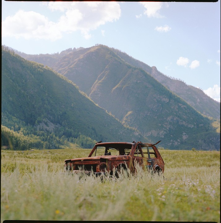 A rusty, abandoned car sits in a field of tall grass with mountains in the background.