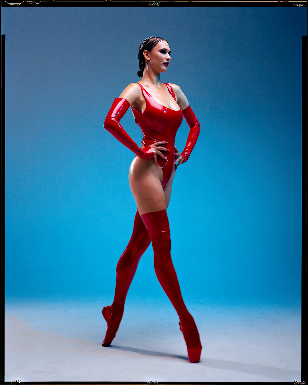 A woman in a red latex bodysuit, gloves, and thigh-high boots stands en pointe with her hands on her hips.