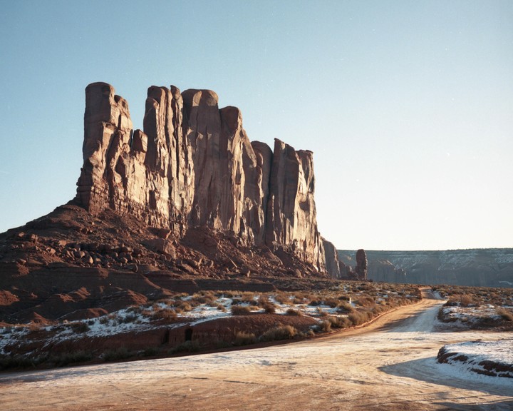 A dirt road leads towards a large rock formation in a desert landscape with patches of snow.