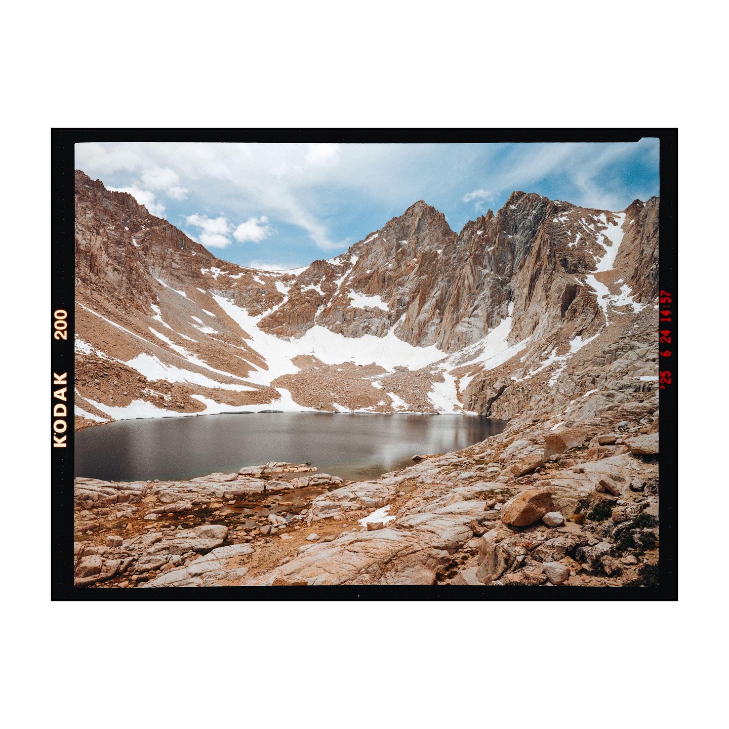 A lake sits at the base of a snow-capped mountain range under a blue sky with wispy clouds.