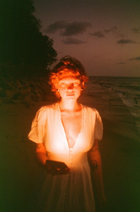 A woman in a white dress stands on a beach at dusk, illuminated by an orange light.