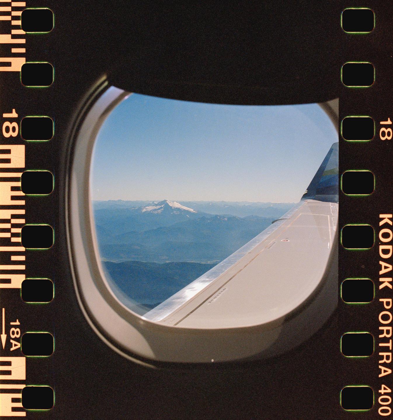 A snow-capped mountain range is visible from an airplane window, with the wing of the plane in the foreground.