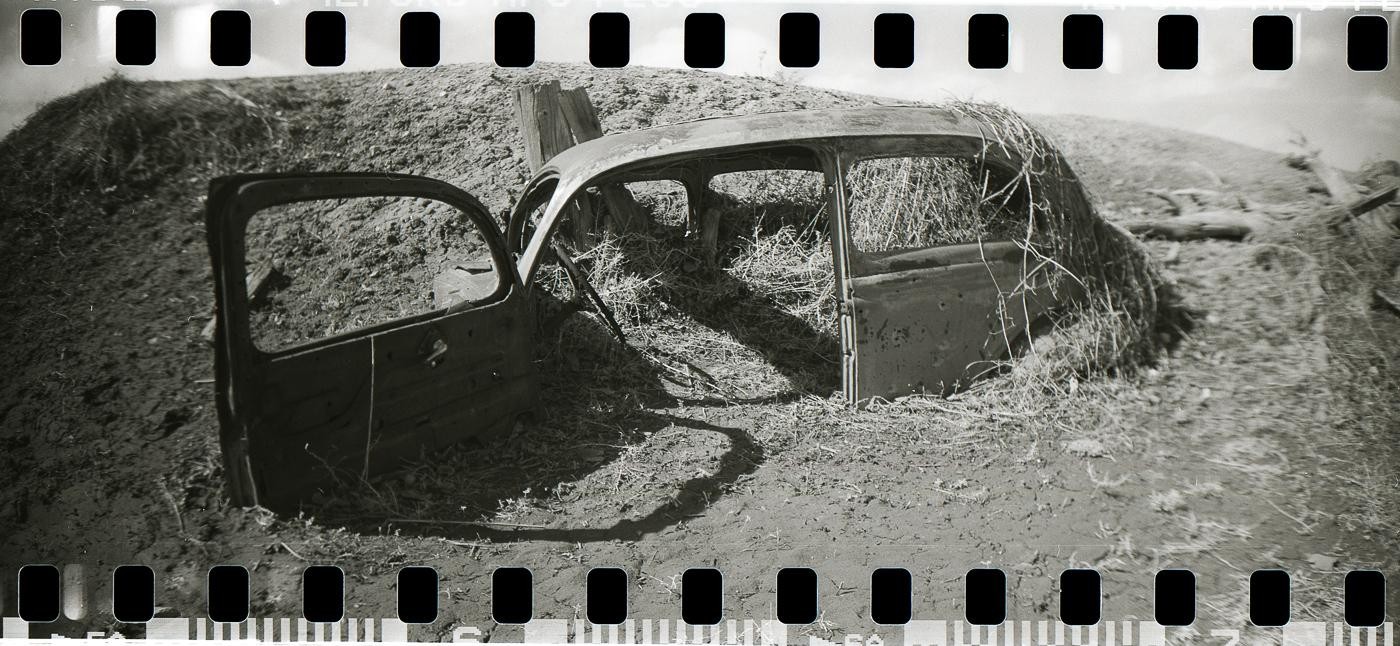 An abandoned car body sits in a field with one door open, surrounded by dry grass and dirt.
