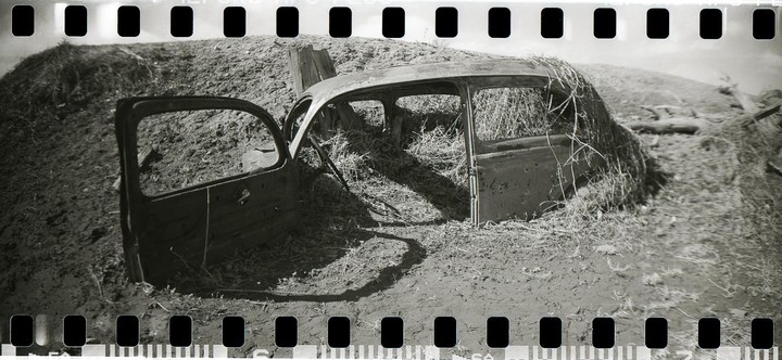 An abandoned car body sits in a field with one door open, surrounded by dry grass and dirt.