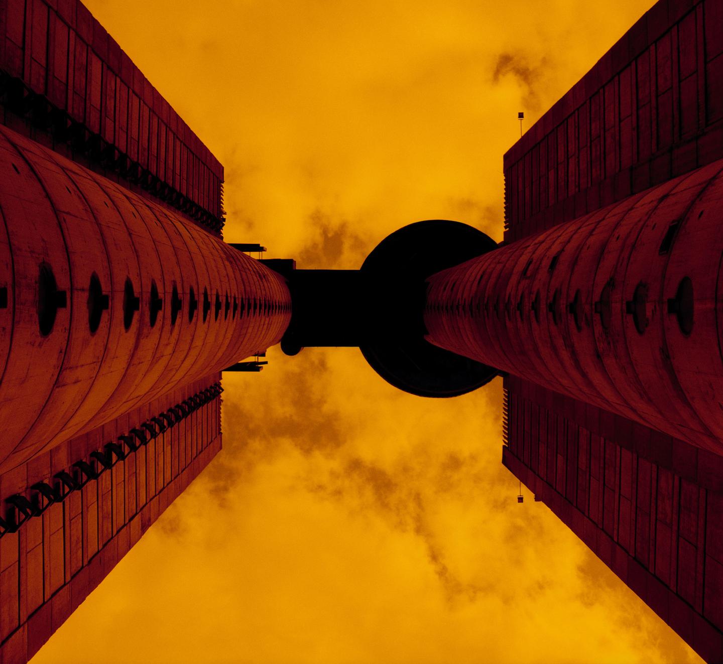 Two tall, cylindrical buildings with dark, circular structures between them are viewed from below against an orange sky.