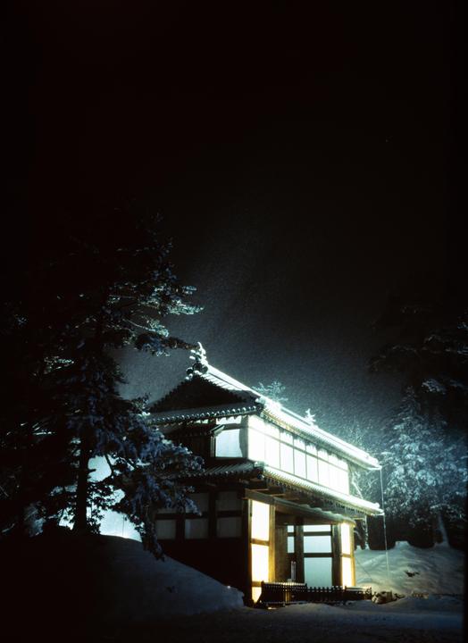 A traditional Japanese building with illuminated windows is surrounded by snow-covered trees at night.