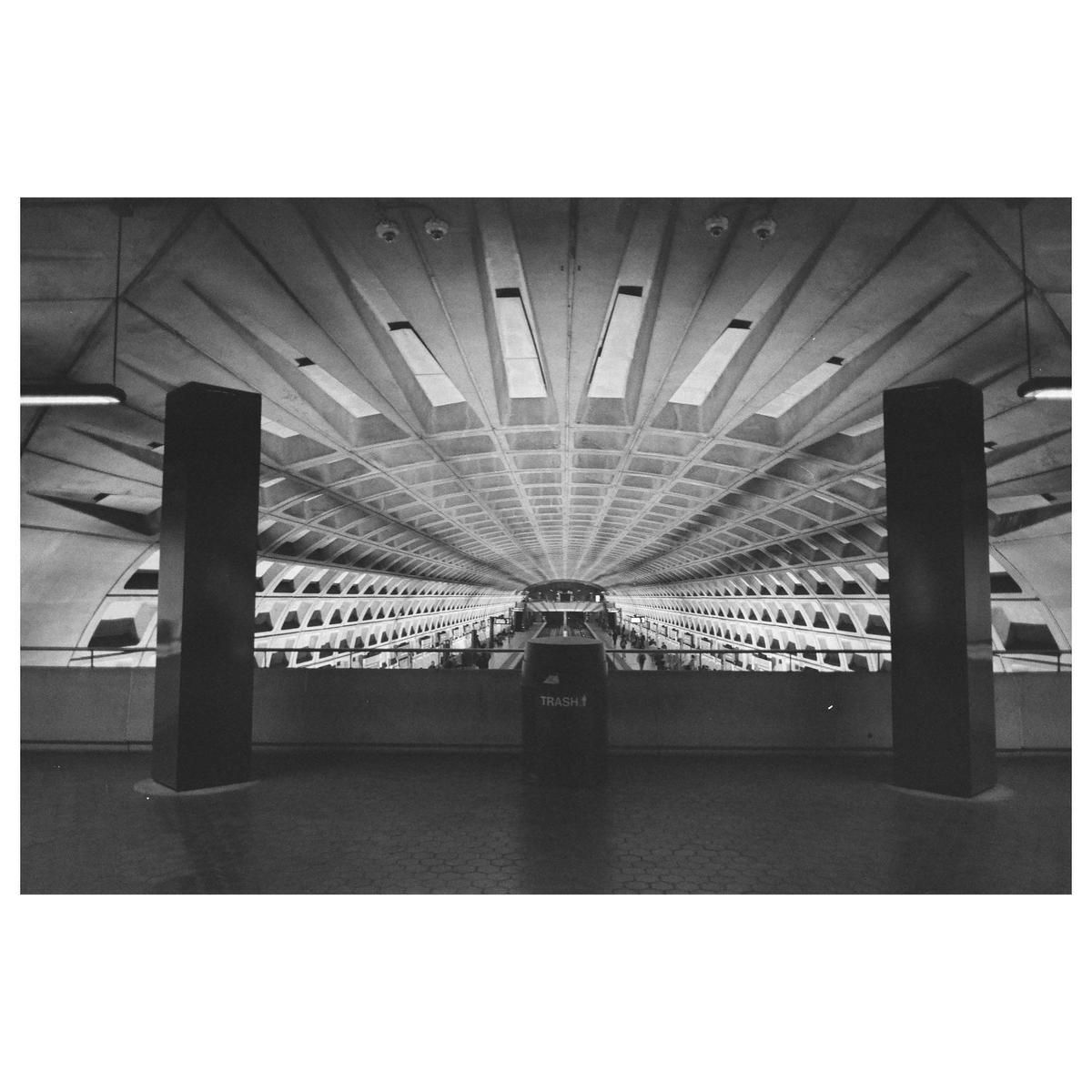 A long, vaulted subway tunnel with a trash can in the foreground is captured in black and white.