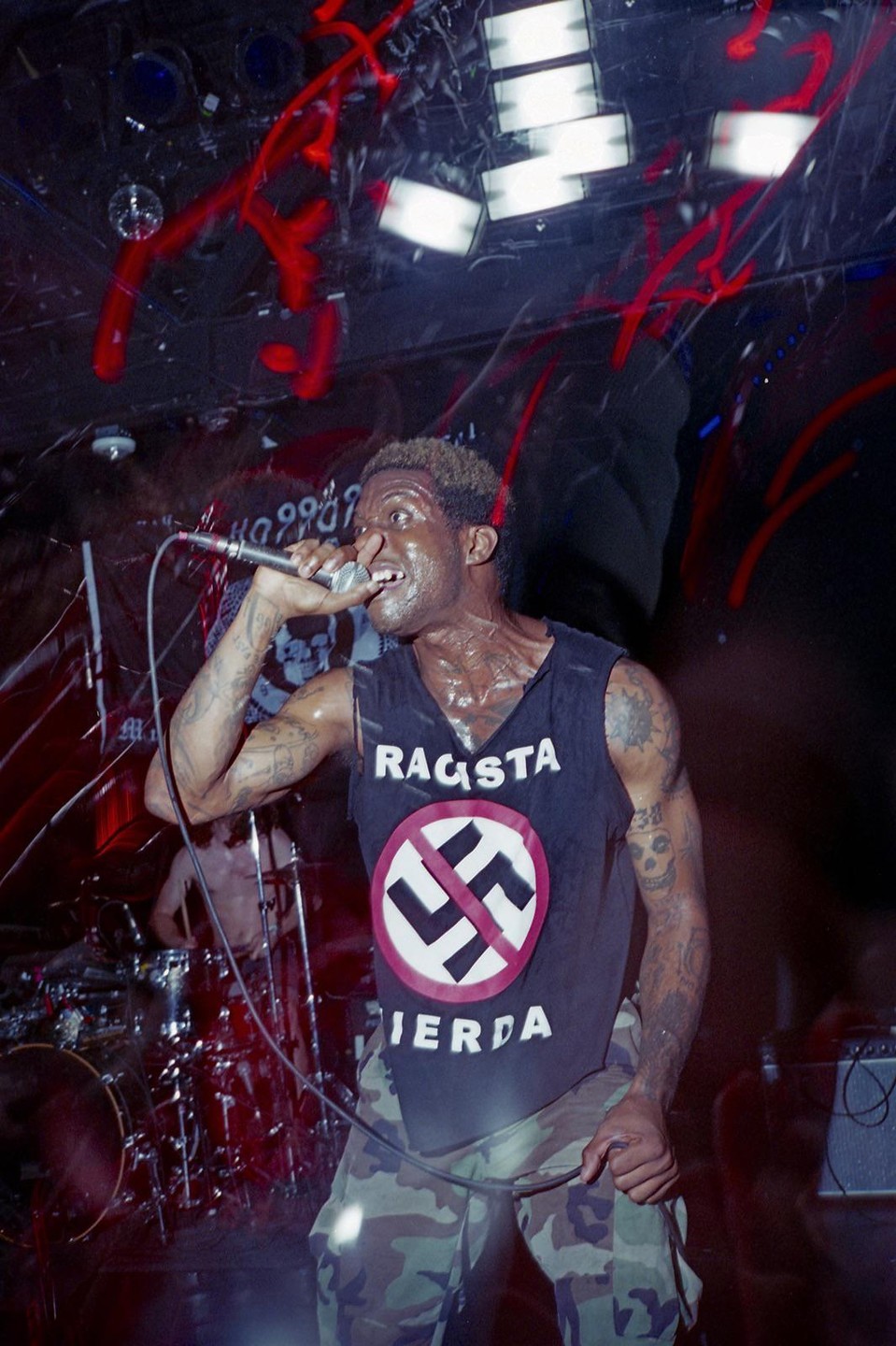 A man with a microphone sings on stage, wearing a black tank top with a red circle and a crossed-out swastika.