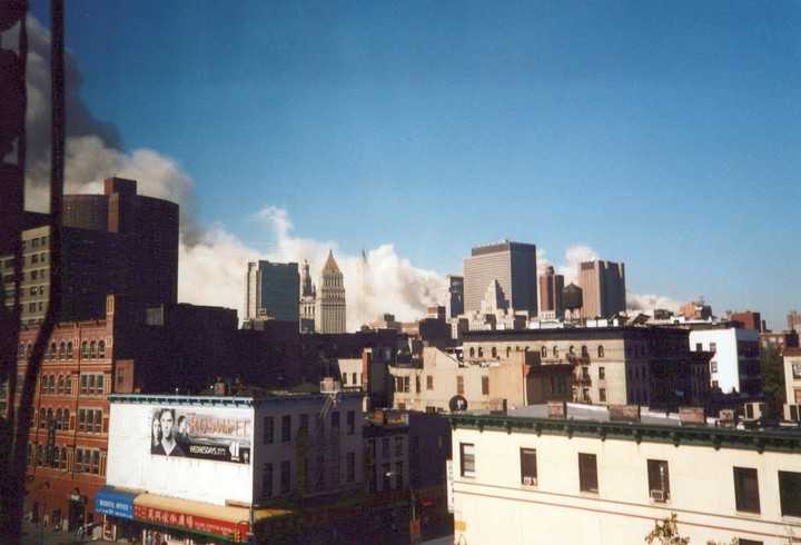 A city skyline is seen with smoke rising from behind buildings under a clear blue sky.