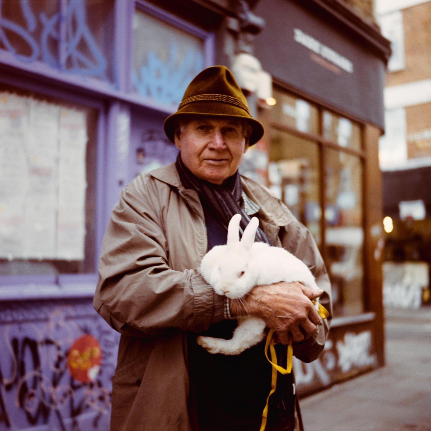 A man wearing a hat and a coat holds a white rabbit with red eyes in his arms on a street.