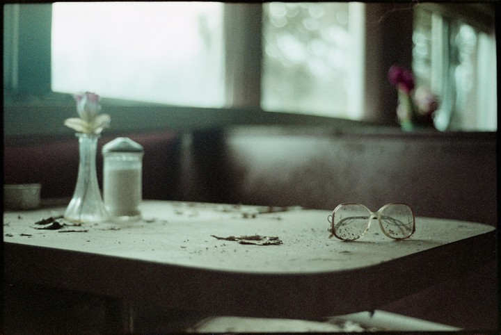 A pair of eyeglasses rests on a dusty table next to a vase with a flower and a sugar dispenser in an abandoned diner.