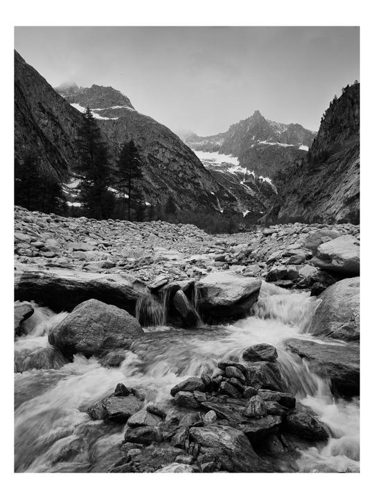 A river flows over rocks in a valley with snow-capped mountains in the background.