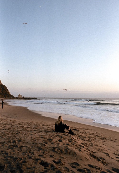 A woman sits on a sandy beach looking out at the ocean with paragliders in the sky and surfers in the water.