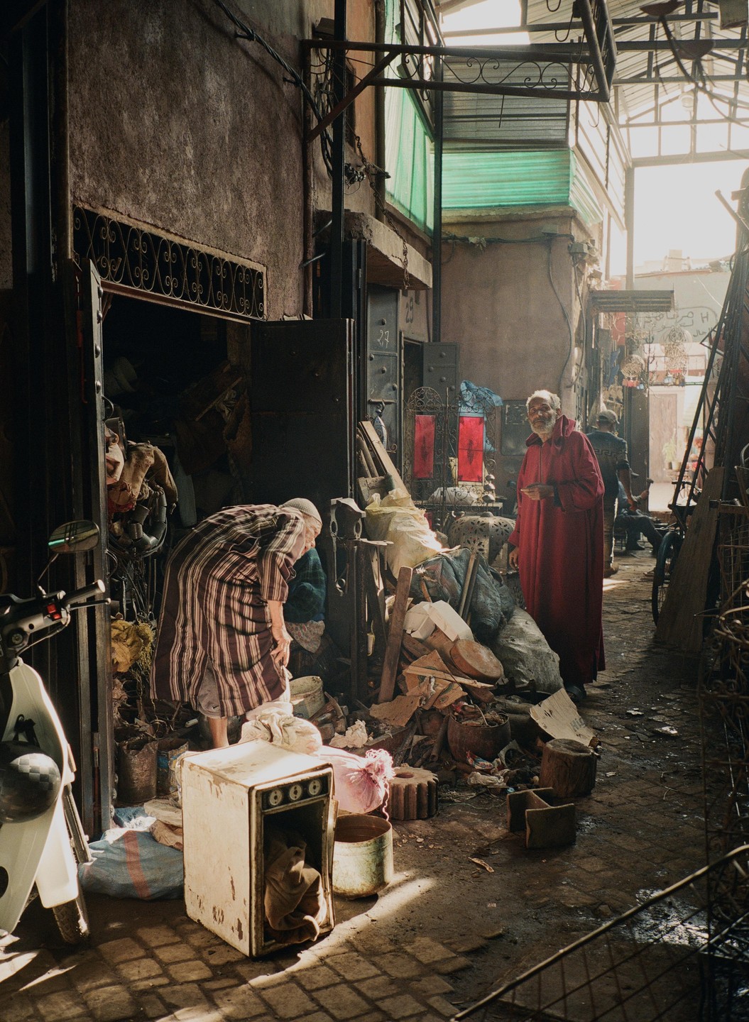 Two men are in a cluttered alleyway with one man wearing a red robe and the other bending over.