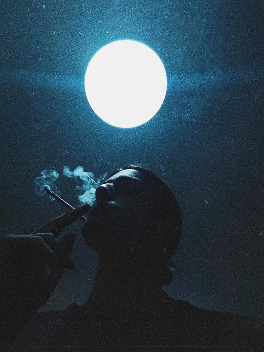 A man in silhouette smokes a cigarette under a bright full moon.