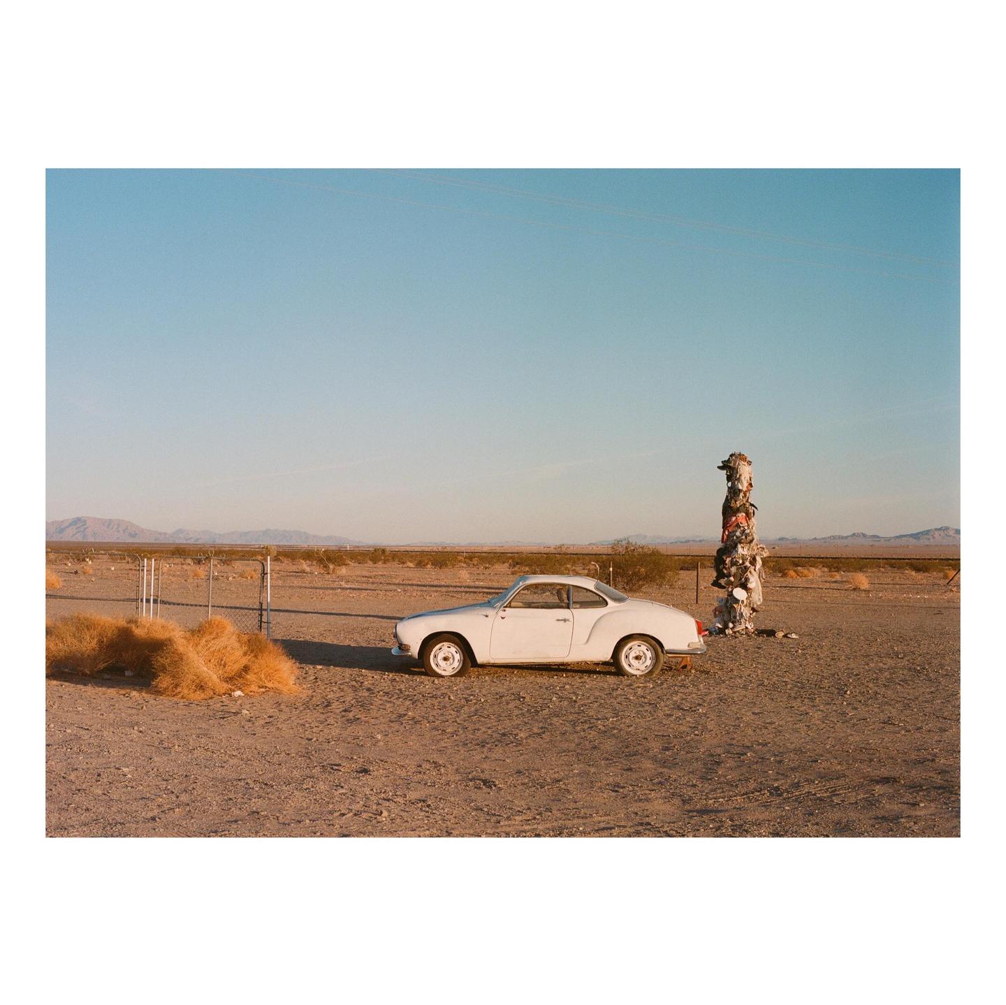 A white car is parked in a desert landscape next to a trash totem and a pile of tumbleweed.