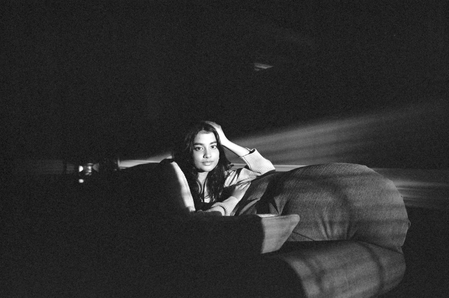A woman reclines on a sofa in a dark room with light rays visible in the background.