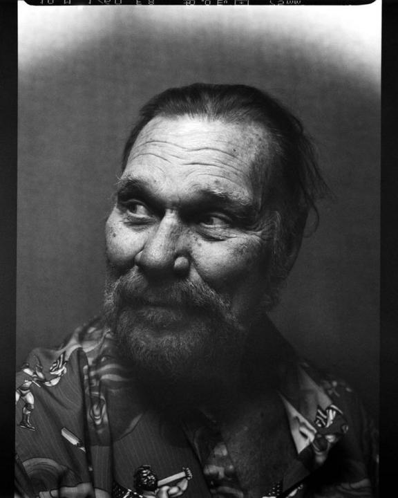 A man with a beard and patterned shirt looks to the side in a black and white portrait.