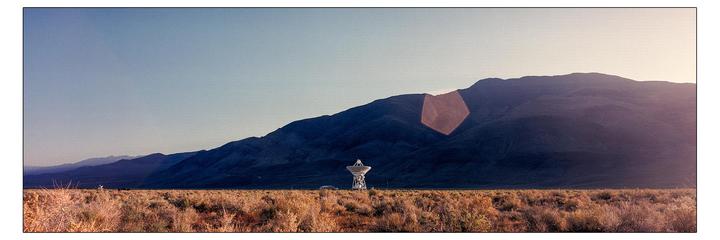 A large satellite dish stands in a field of dry brush with mountains in the background under a clear sky.