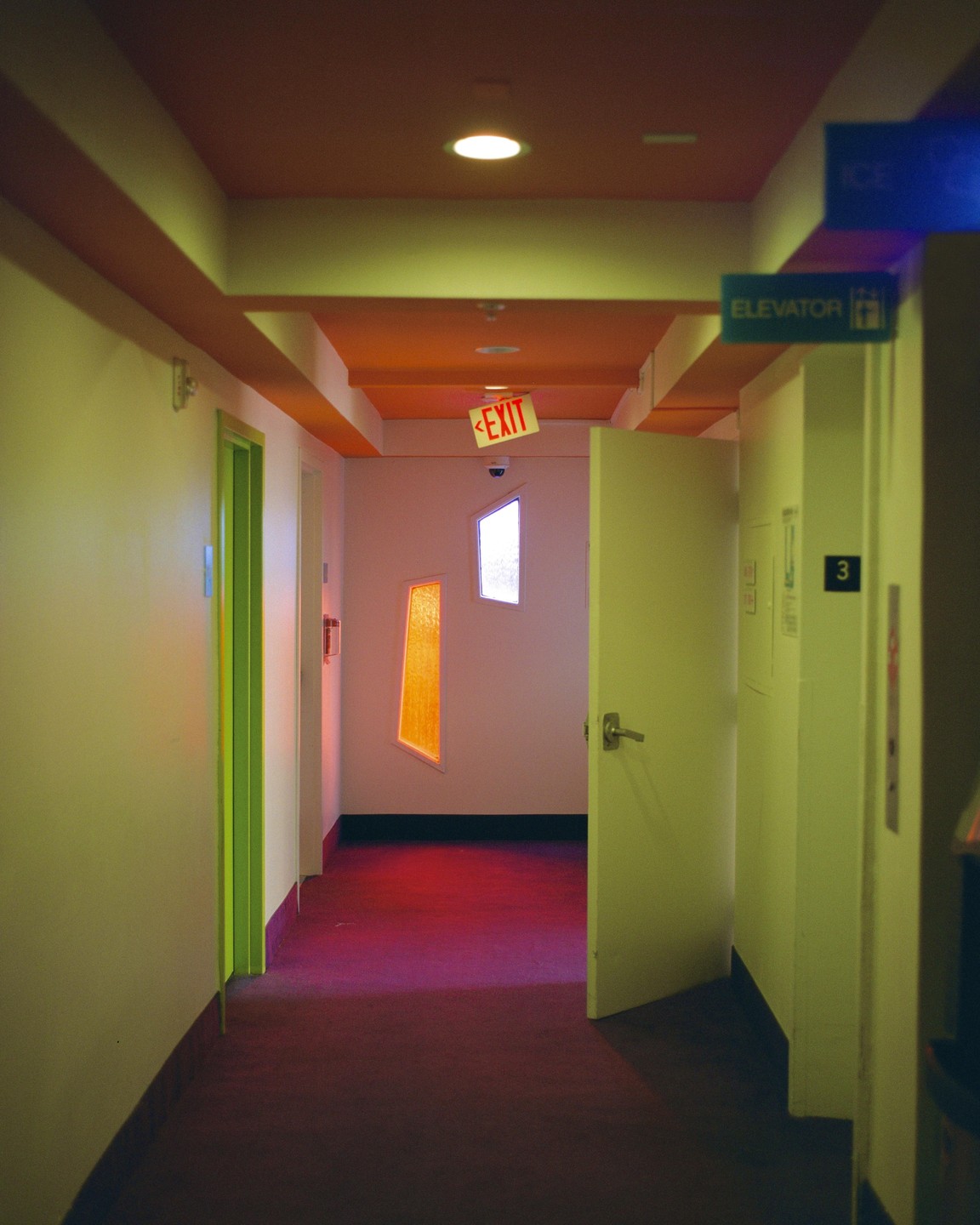 A hotel hallway with an open door, an exit sign, and colorful windows.