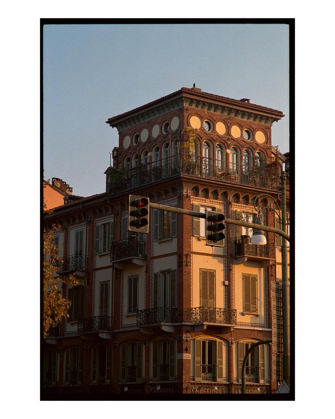 A brick building with balconies and a decorative top floor is illuminated by golden hour light, with traffic lights in the foreground.